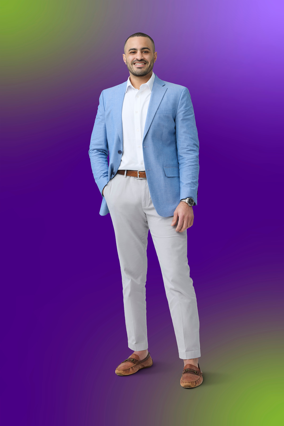 Man in a light blue blazer, white shirt, and beige pants standing confidently against a vibrant purple and green gradient background, smiling with one hand in his pocket.Man in a light blue blazer, white shirt, and beige pants standing confidently against a vibrant purple and green gradient background, smiling with one hand in his pocket.