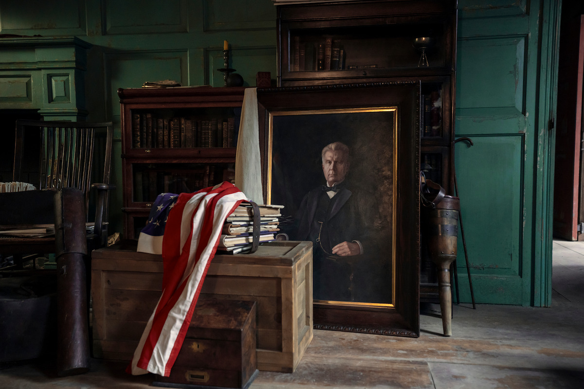 A vintage study with bookshelves, a large framed portrait of a man, wooden crates, stacked books, and an American flag draped over the boxes, all in a dimly lit room with green paneled walls and rustic decor.