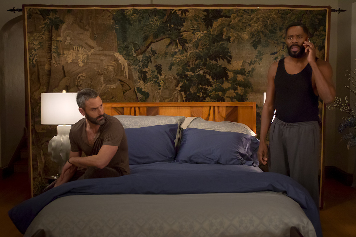 Two men in a bedroom, one sitting on a neatly made bed with blue bedding, the other standing and talking on the phone, both appearing serious. The room has warm lighting and a decorative tapestry hanging behind the bed.