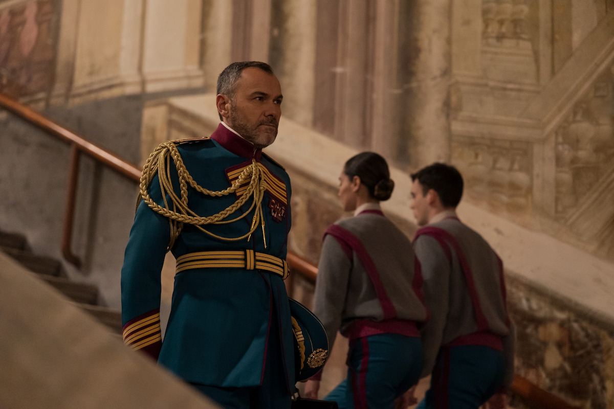 Homem de meia idade e cabelos grisalhos veste uniforme militar e desce a escada de um salão