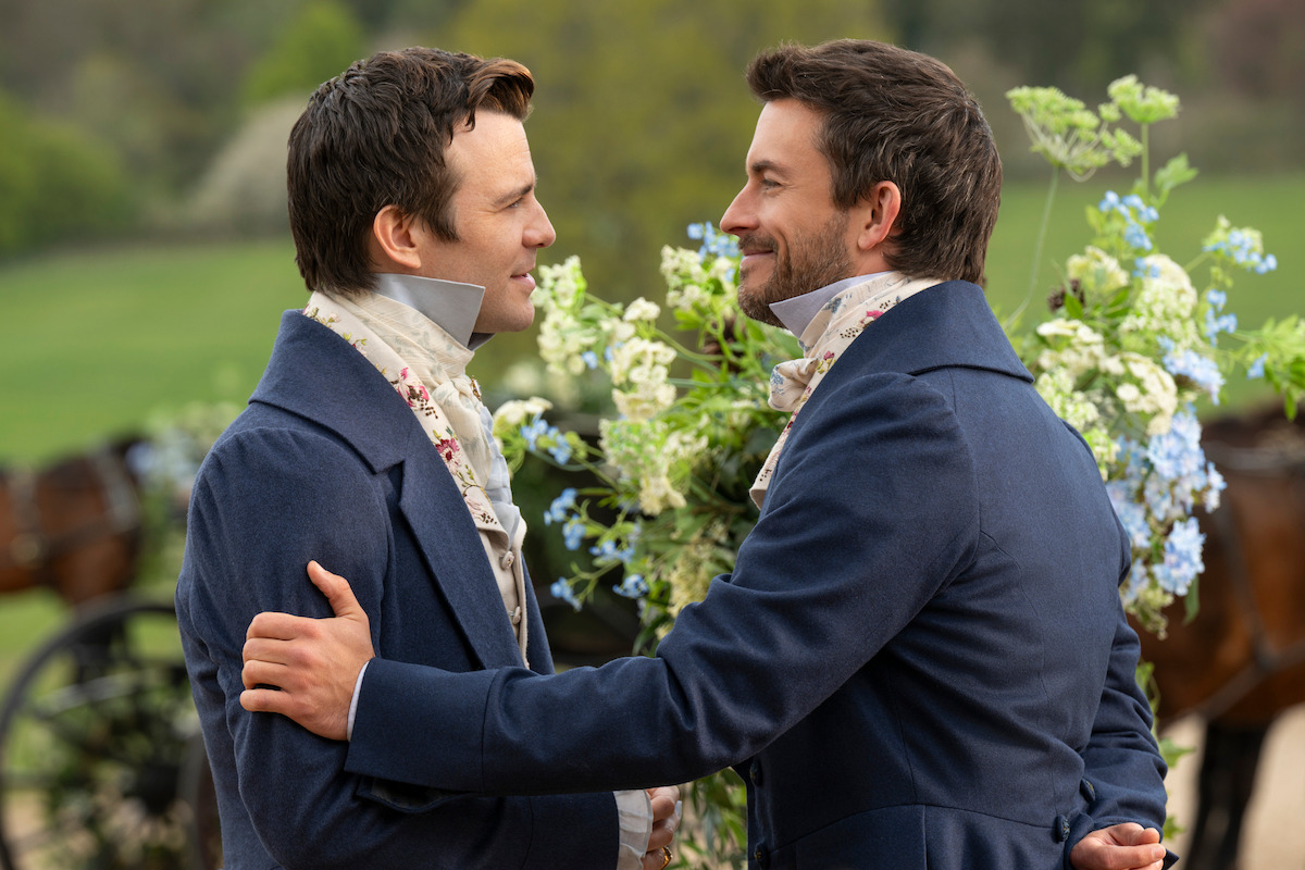 Two men in Regency-era clothing embrace outdoors, smiling at each other. The background features flowering bushes and greenery, suggesting a garden or countryside setting with a carriage partially visible behind them.