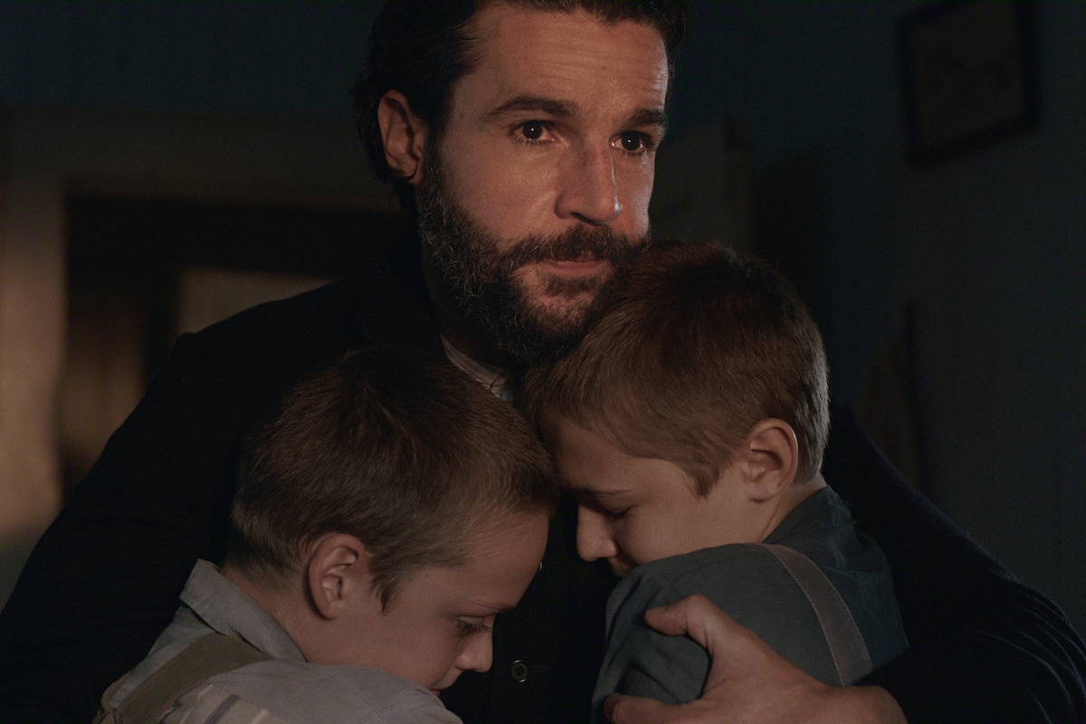 A bearded man in a dimly lit room hugs two young boys close to his chest, providing comfort. All three have serious, emotional expressions, suggesting a moment of sadness or reassurance in a home setting.