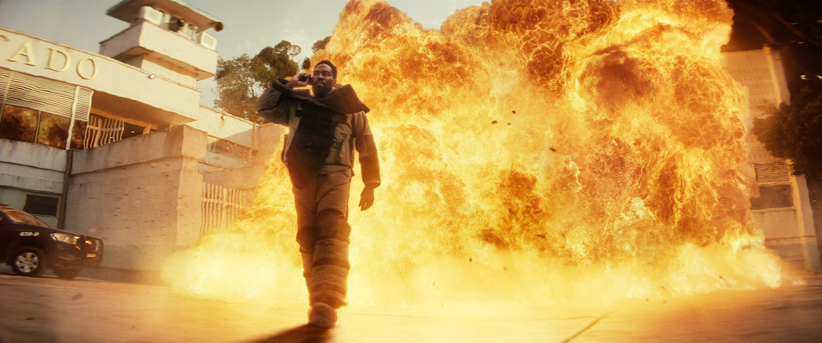 A person in protective gear confidently walks away from a massive explosion outside a building in an urban environment, with fire and smoke dominating the background.