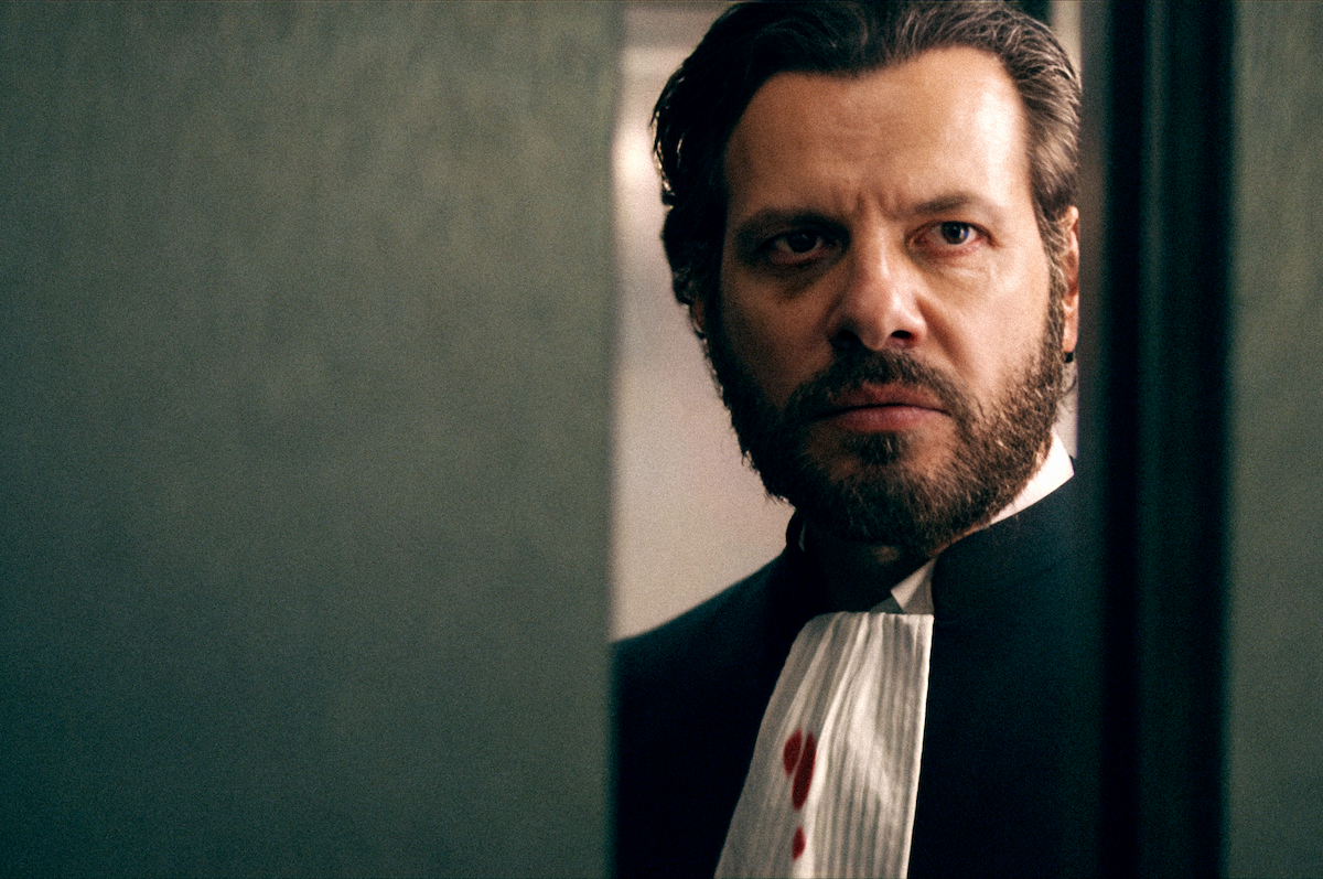 A serious man with a beard and formal attire stands in a dimly lit room, partly viewed through a doorway, with blood stains on his white collar, creating a tense, dramatic atmosphere.