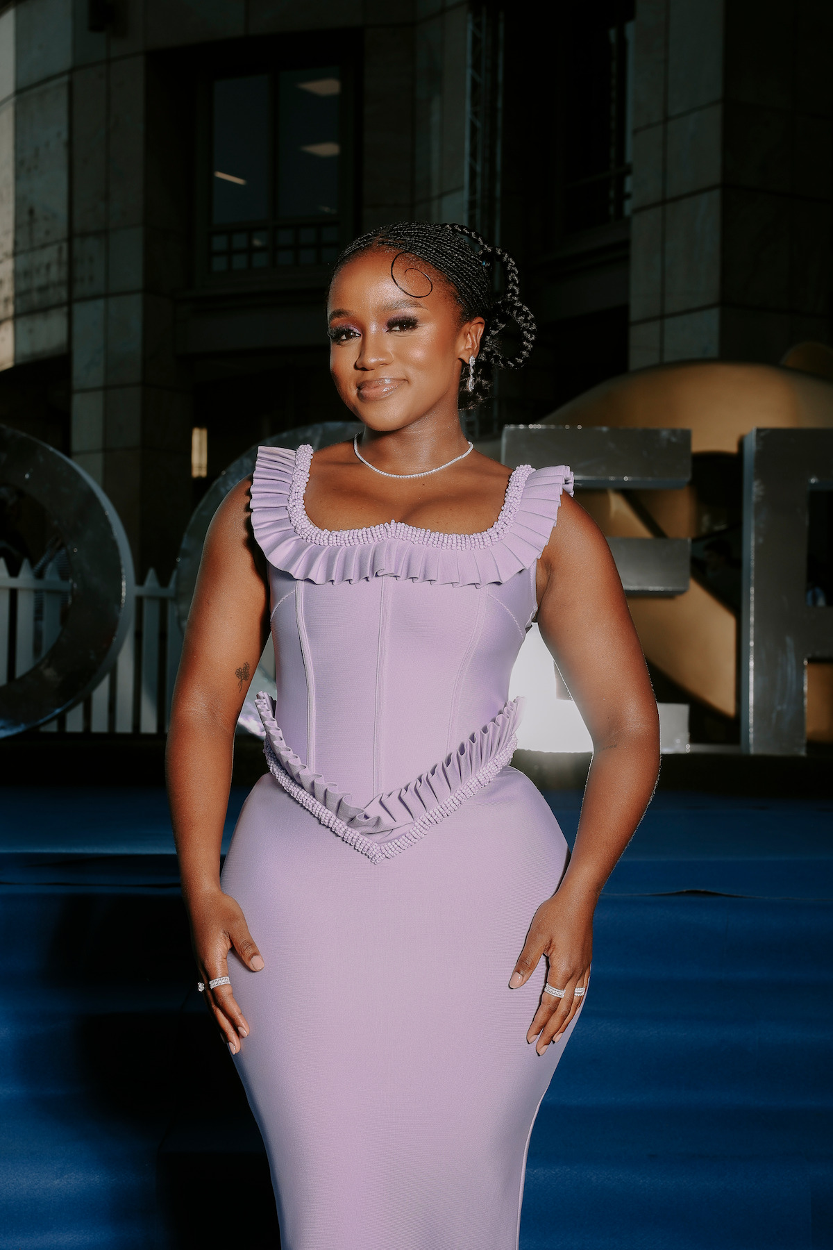 Woman in elegant lavender dress with ruffled details posing on blue carpet at a formal event, outdoor setting with modern architectural background and decorative elements.