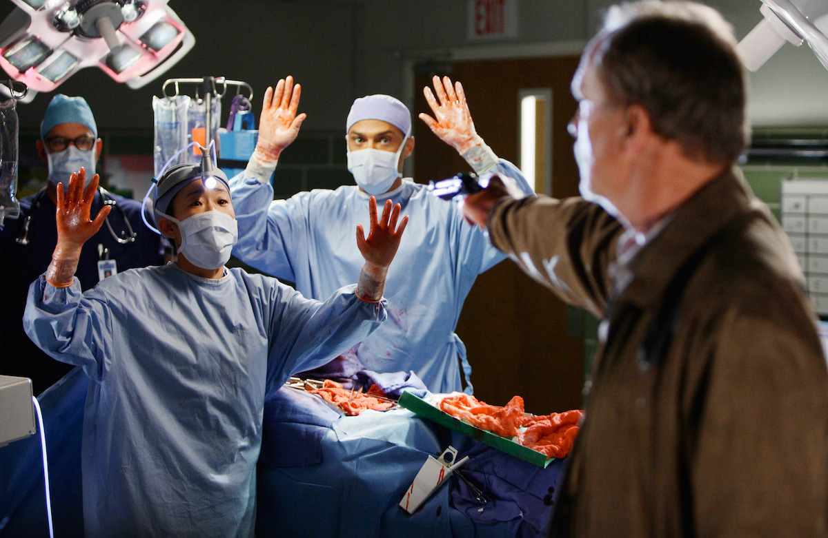 a group of doctors performing surgery hold their hands up while a man holds a gun on them. 