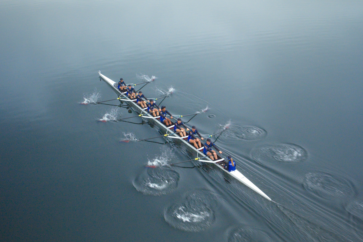 Aerial view of a rowing team in a long boat moving in sync across calm, reflective water, creating ripples as they row together in a misty, tranquil outdoor environment.