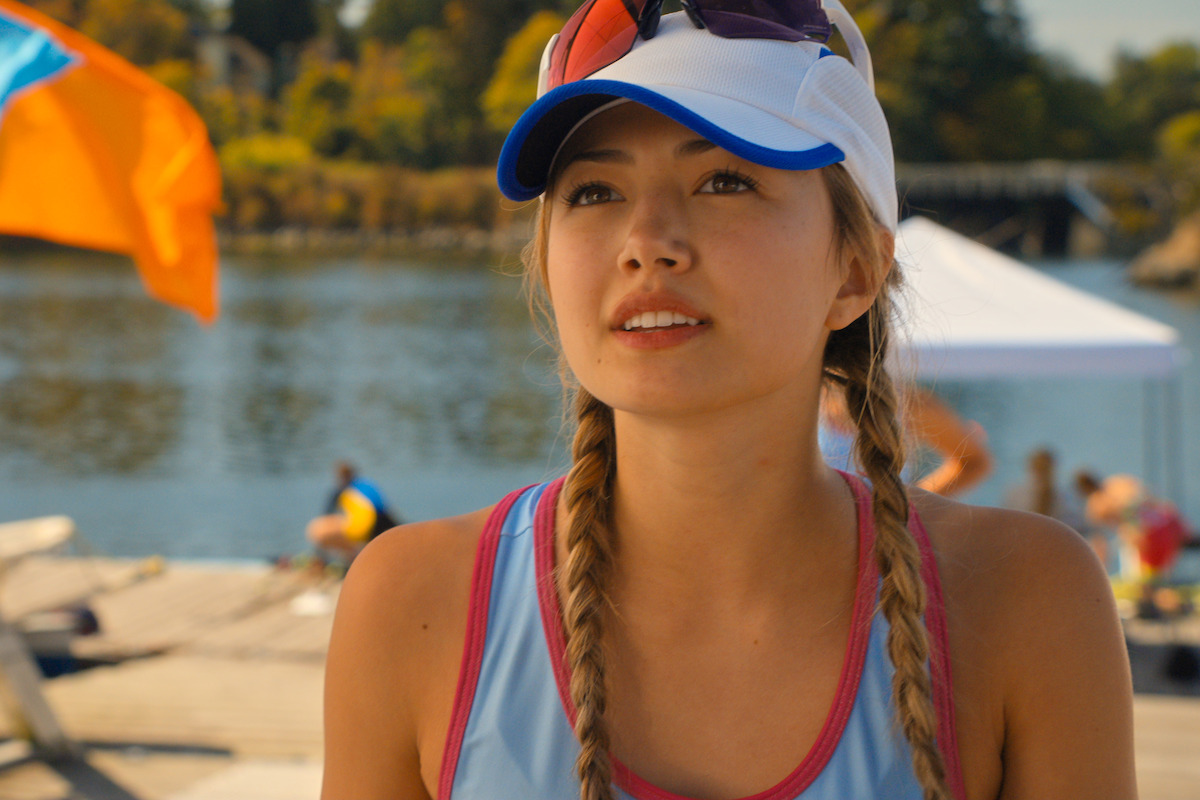 Young woman with braided hair, wearing a cap and sunglasses, stands outdoors by a river, dock, and trees, in bright daylight, with people and colorful flags in the background, suggesting a warm, recreational setting.