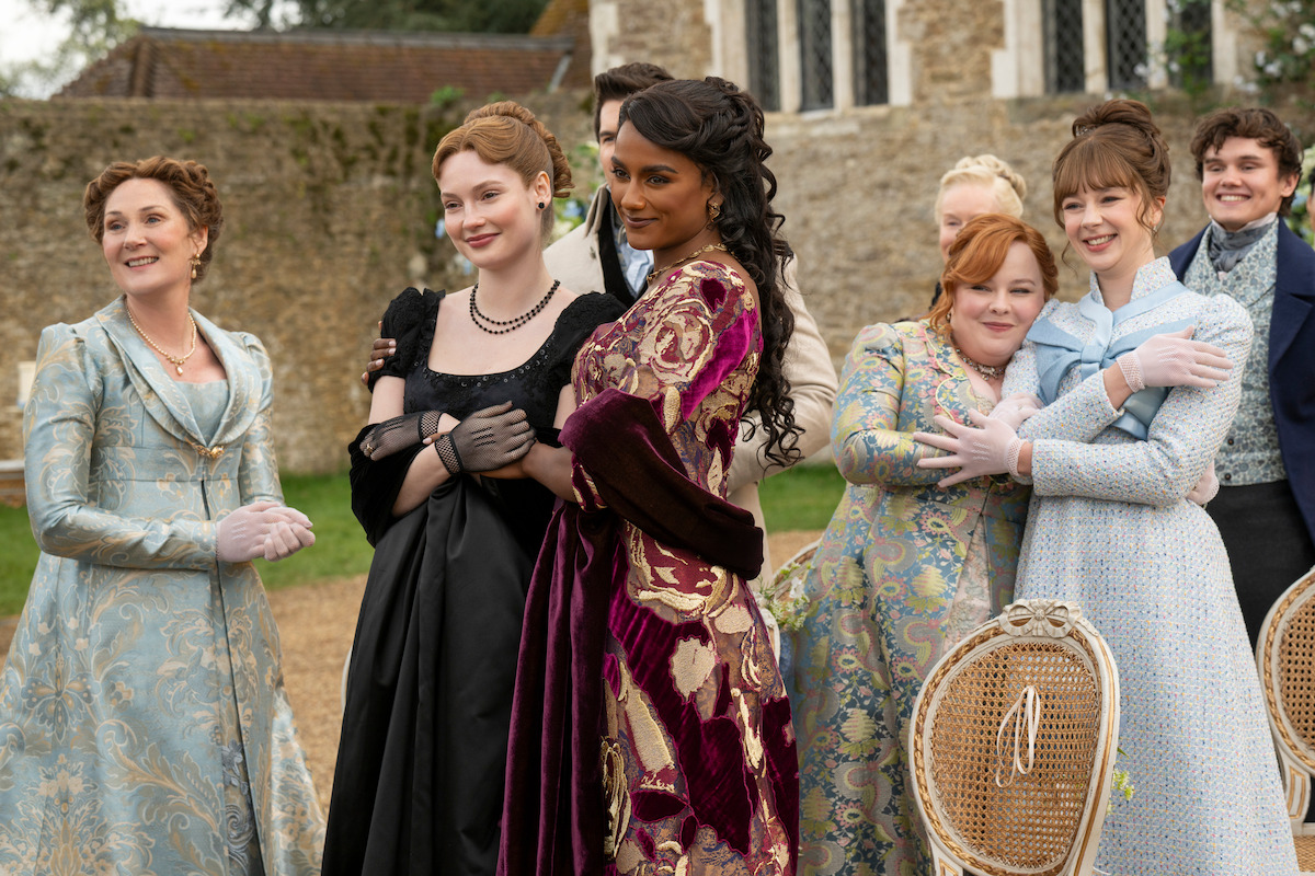 A group of people in elaborate period costumes stand outdoors in a garden or courtyard, smiling and posing together in front of old stone buildings, with chairs visible in the foreground.