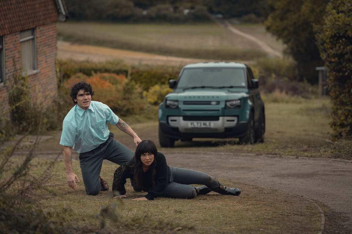 A man and woman appear startled as they sit on the ground outside near a brick house, with a green SUV parked on a rural driveway and fields in the background on a cloudy day.