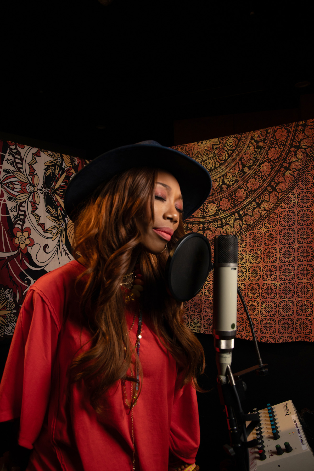 Woman in a hat and red shirt singing into a microphone in a music studio with vibrant, patterned wall art and audio equipment visible in the background, creating an artistic and creative atmosphere.