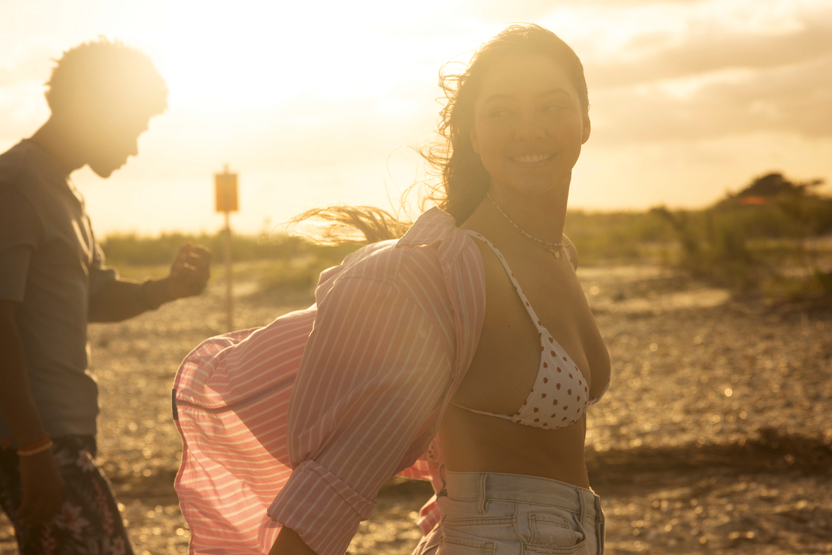 Smiling woman in a polka-dot bikini top and open shirt walking on a sunny beach at sunset, with another person in the background, warm natural light and relaxed atmosphere.