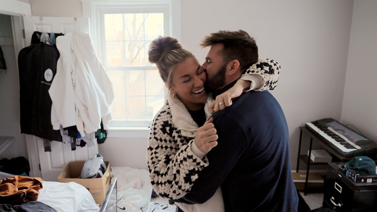 A smiling couple embraces in a cozy, cluttered bedroom with clothes scattered, a laundry basket, window, and a keyboard on a desk in the background.