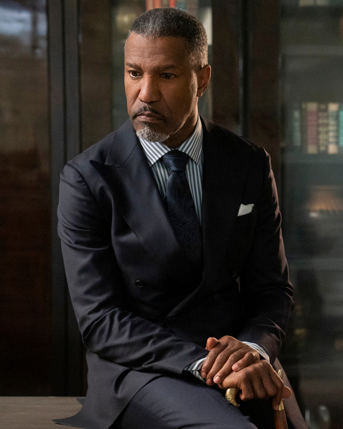 Man in a dark suit with a striped shirt and tie, sitting indoors holding a cane, with shelves and books visible in the background, looks serious and thoughtful in a formal office or study setting.