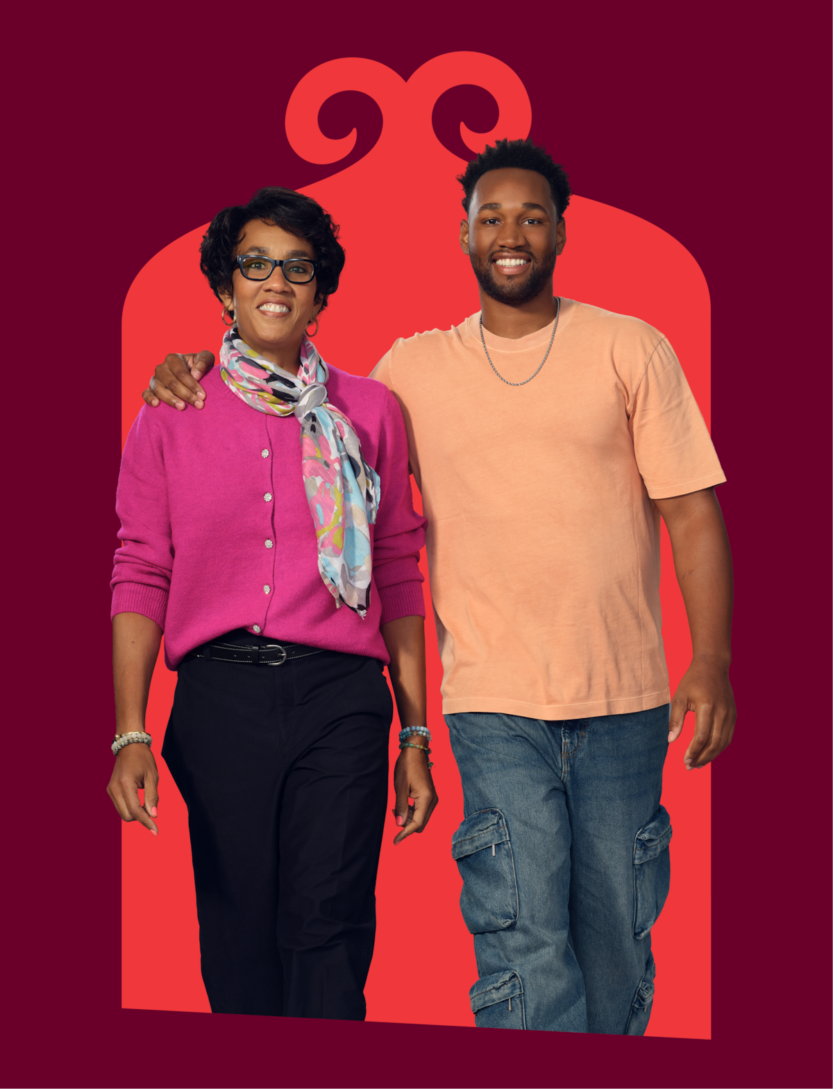 Two people standing together in front of a decorative, red archway on a burgundy background, one wearing a pink cardigan and scarf, the other in a peach T-shirt and jeans, both smiling and looking at the camera.