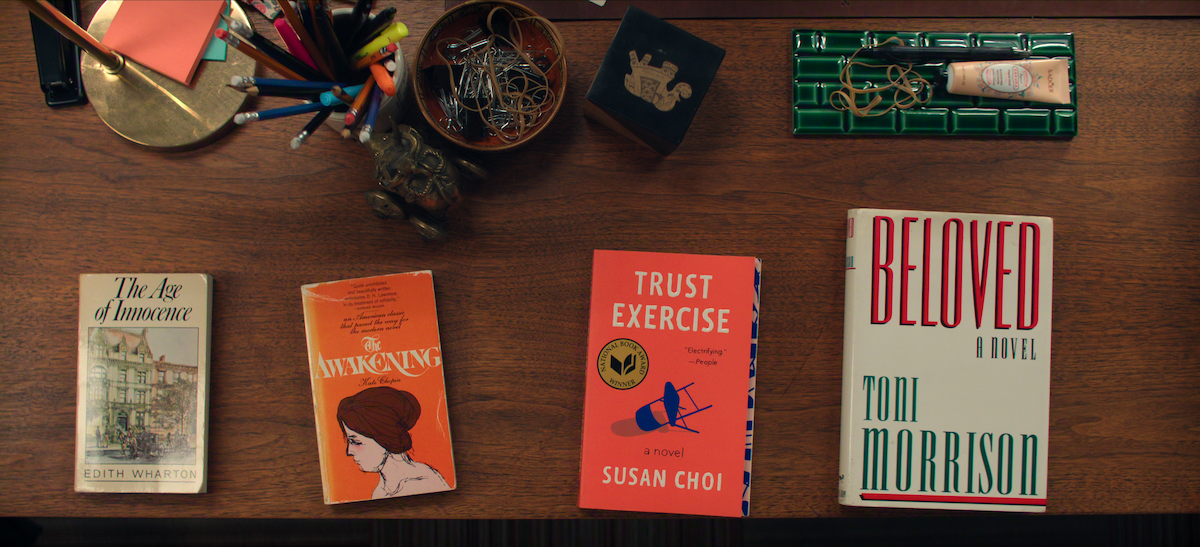 Rachel Weisz as The Protagonist standing in front of classroom chalkboard, holding book and smiling, with desk in foreground holding papers and a briefcase, academic setting.