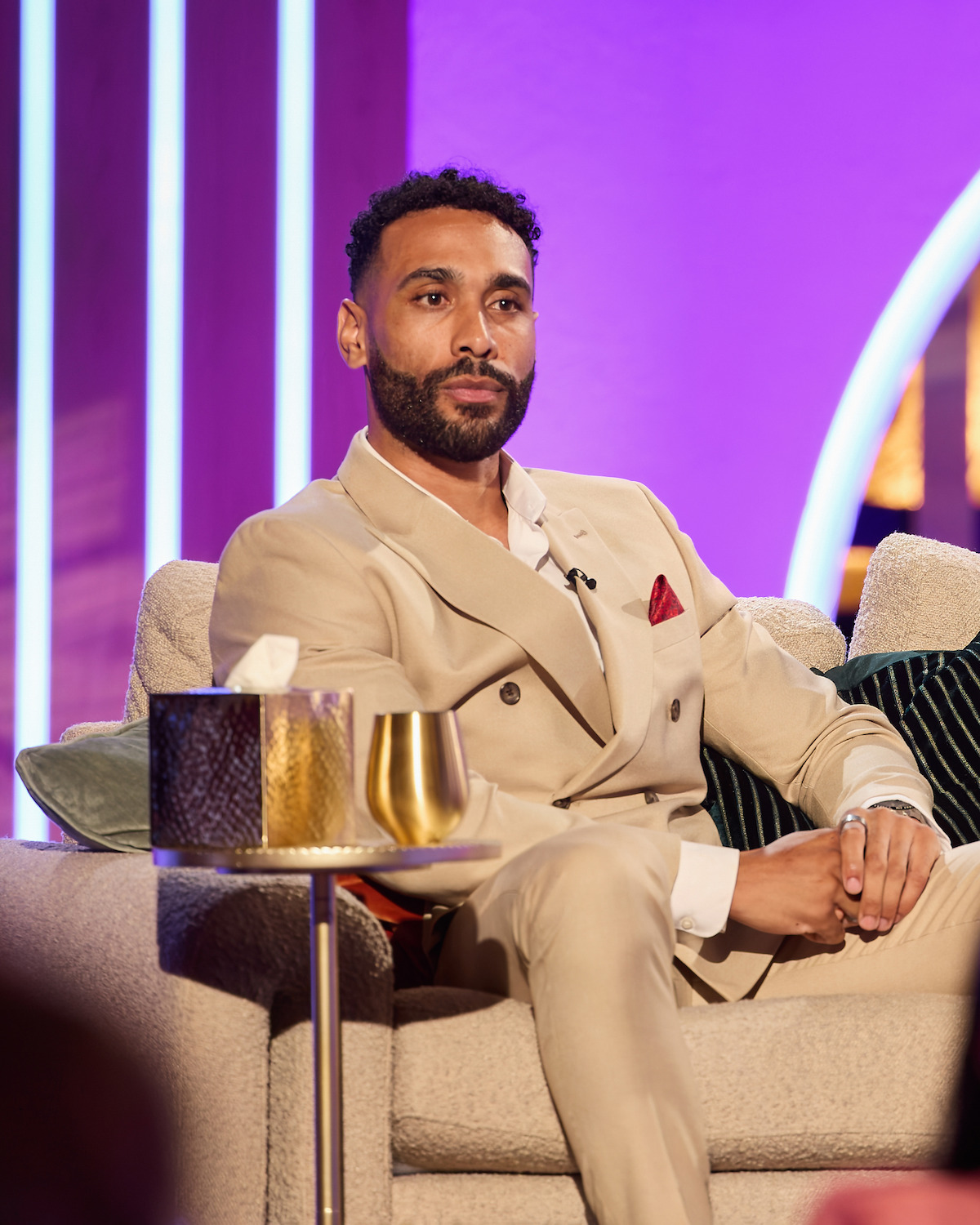 Man in a beige suit sitting on a sofa in a modern, brightly lit studio with purple lighting, a side table with a gold cup and tissue box in the foreground, and decorative pillows around him.