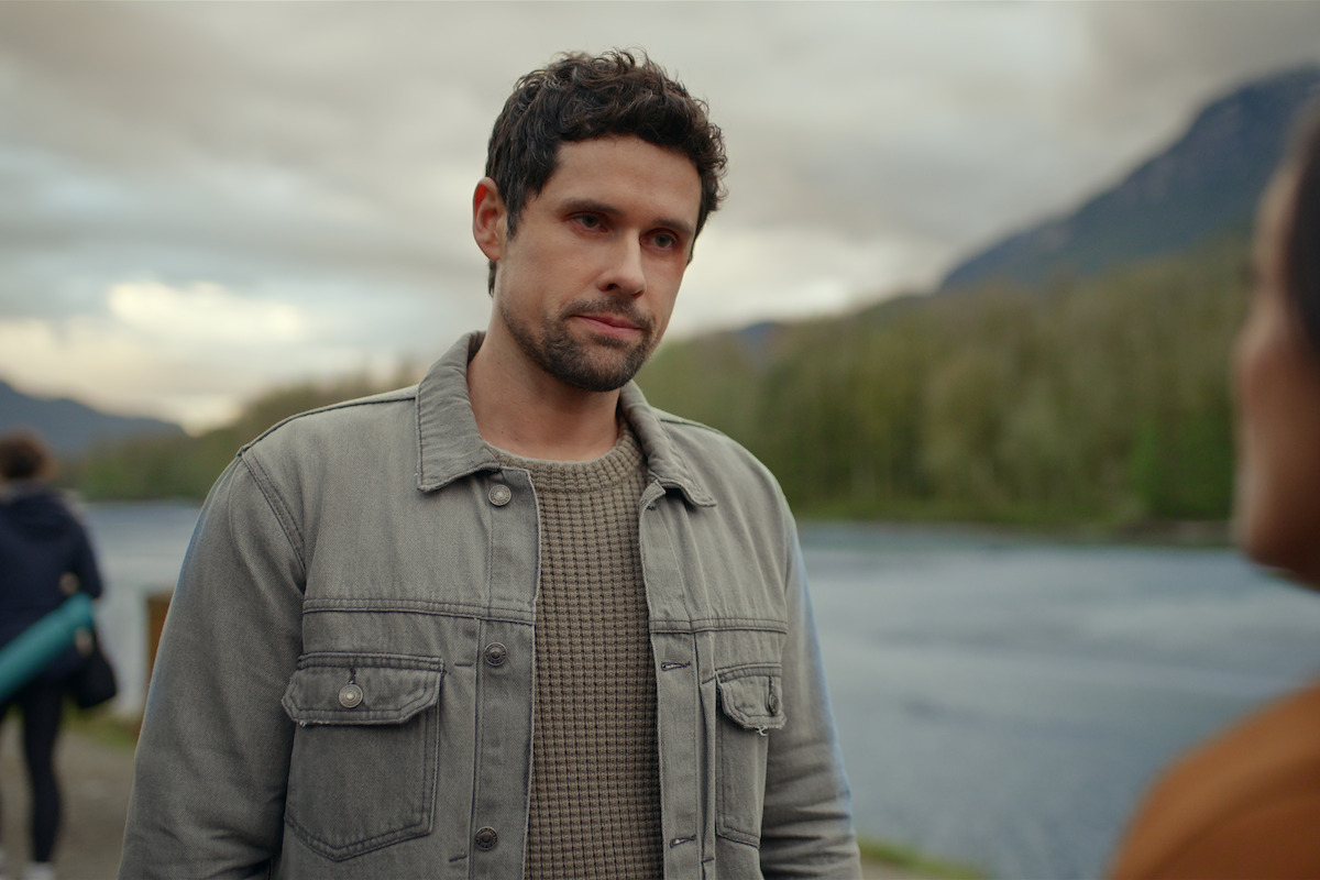A man in a gray jacket stands outdoors near a river, with blurred trees and mountains in the background and cloudy sky above. Another person is in the foreground, and someone is walking away in the distance.