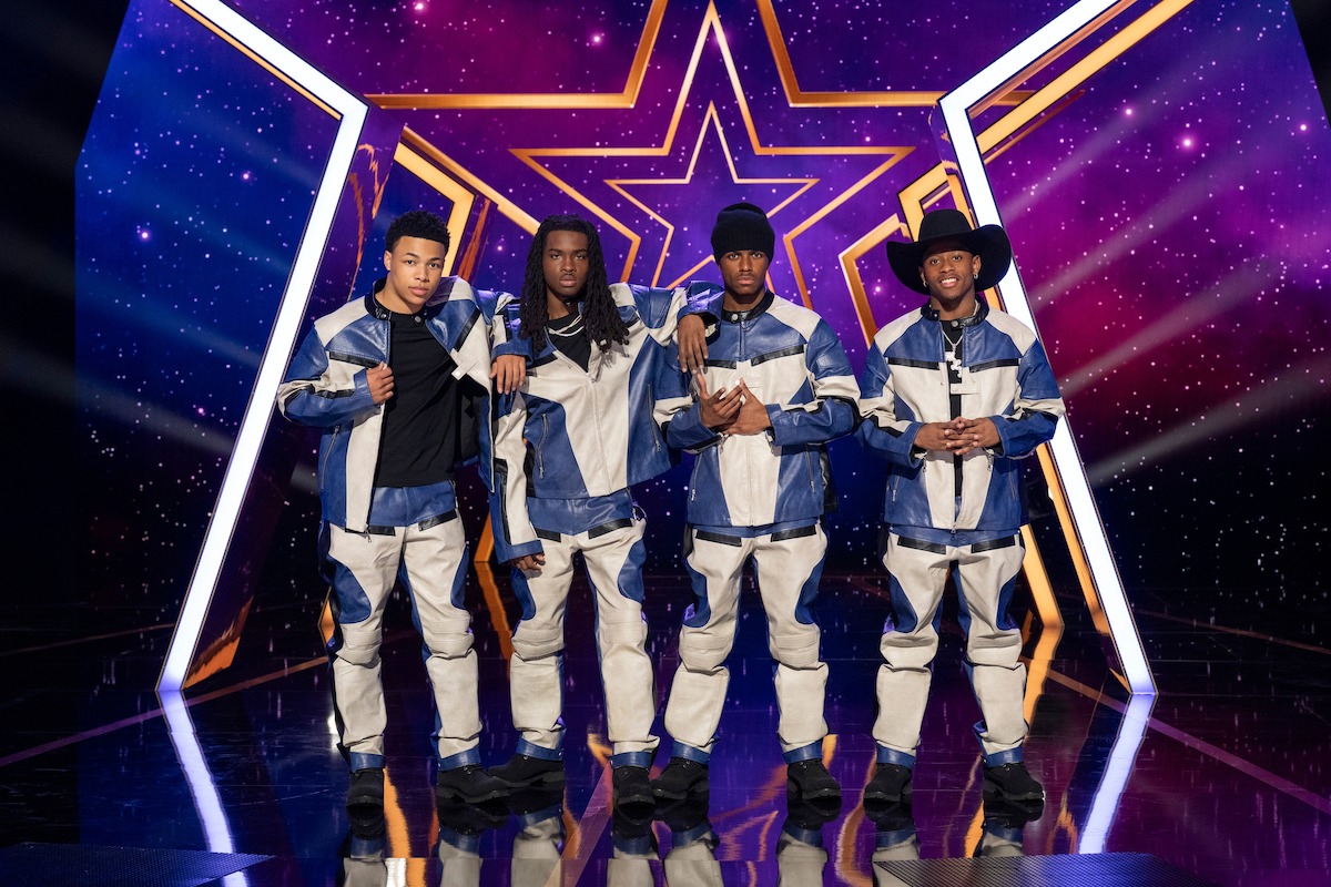 2BYG con trajes azules y blancos a juego están juntos en un escenario brillante con un fondo estrellado colorido y un gran símbolo de estrella detrás de ellos, lo que sugiere un escenario de actuación o espectáculo de talentos.