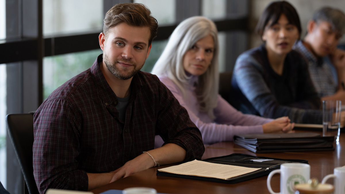 A group of people sit around a conference table in a meeting room with large windows, notebooks and coffee mugs are on the table, while Leo Woodall as Vladimir in the foreground is attentively listening.A group of people sit around a conference table in a meeting room with large windows, notebooks and coffee mugs are on the table, while Leo Woodall as Vladimir in the foreground is attentively listening.