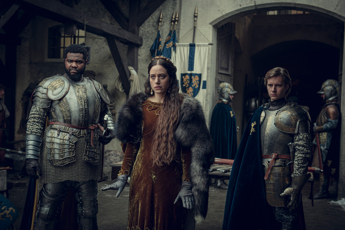 Three medieval figures—two armored knights and a noblewoman in a fur-trimmed gown—stand in a castle courtyard with banners and other knights in the background, creating a tense, dramatic atmosphere.