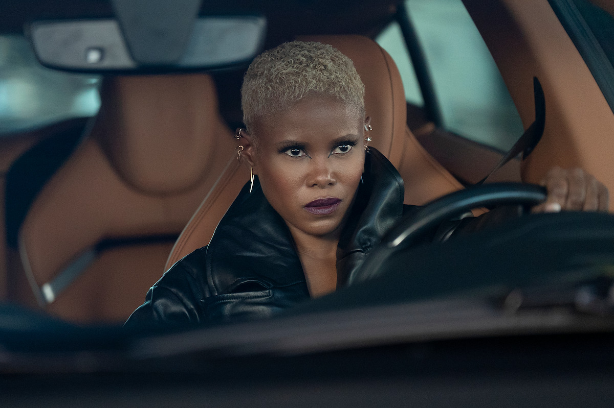 Person with short blonde hair and dark lipstick, wearing a black jacket, sits in the driver’s seat of a luxury car with tan interior, looking focused and intense as they drive.