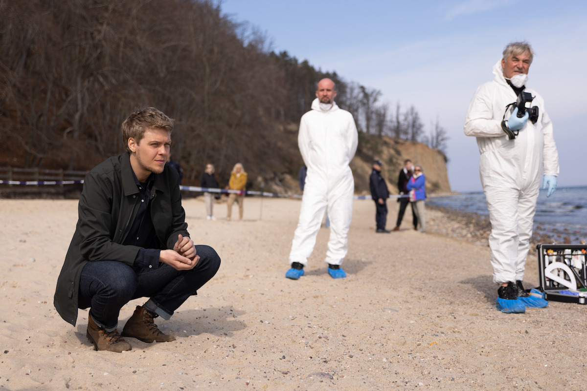 Oprócz gapiów i innych śledczych Jakub Gierszał w roli Leopolda Bilskiego bada miejsce zbrodni na plaży na kadrze z filmu „Kolory zła: czerwony”.