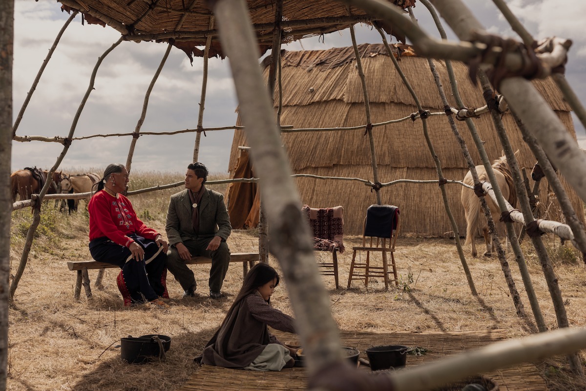 Two adults sit talking under a wooden shelter frame in a grassy field, with a child playing nearby. Behind them are traditional huts and horses, suggesting a rural or indigenous setting. The sky is cloudy.
