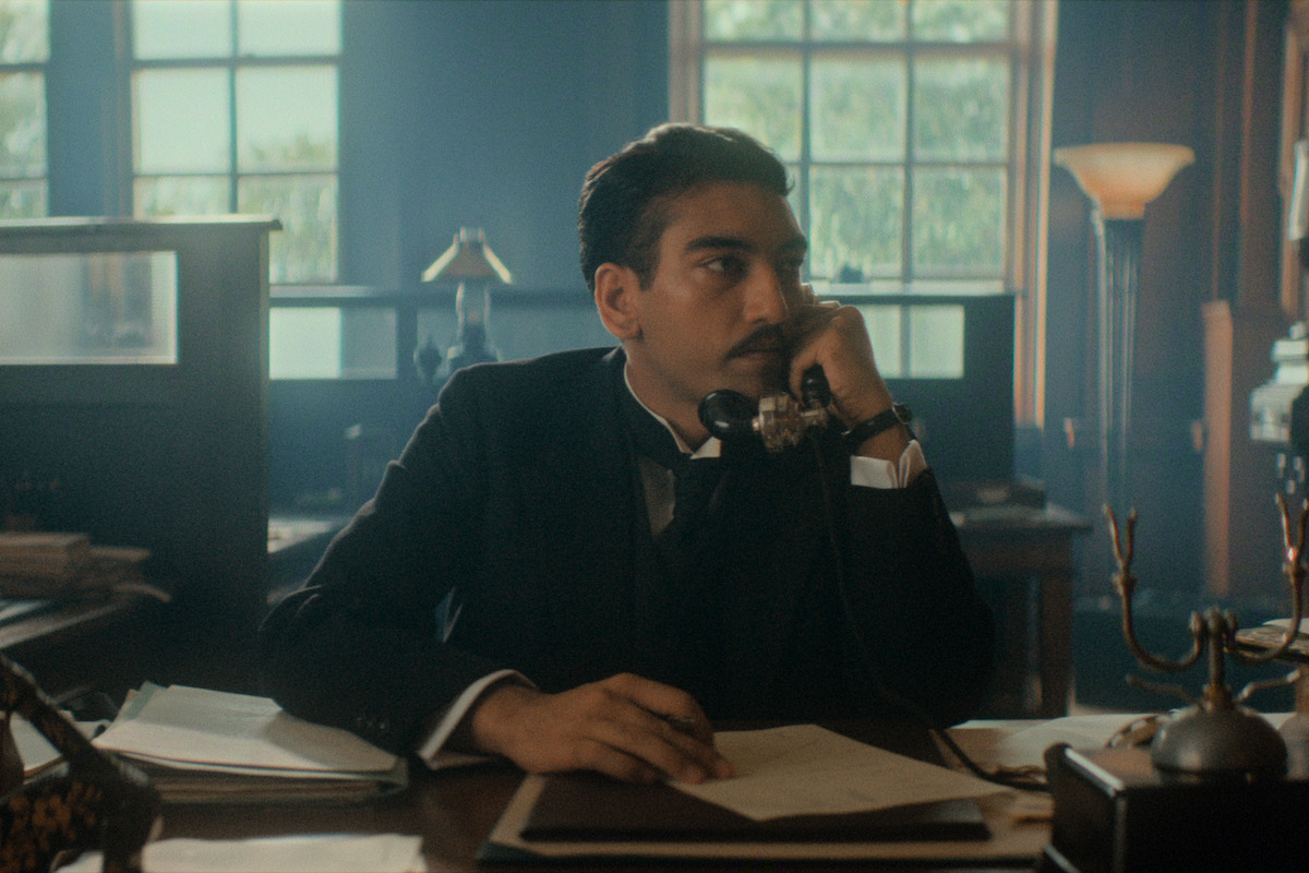 A man in a suit sits at a wooden desk in an old-fashioned office, talking on a rotary phone, with papers, a lamp, and vintage decor around him, and large windows letting in natural light.