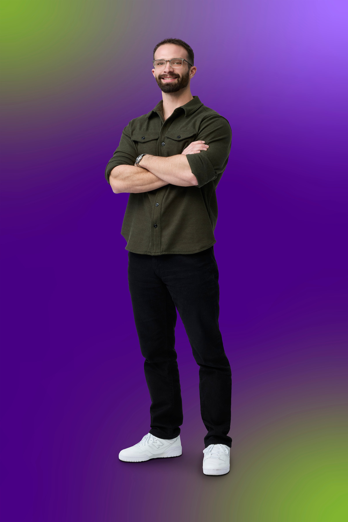 Man with glasses, beard, and short hair stands confidently with arms crossed, wearing a green shirt, black pants, and white sneakers, on a vibrant purple and green gradient background.