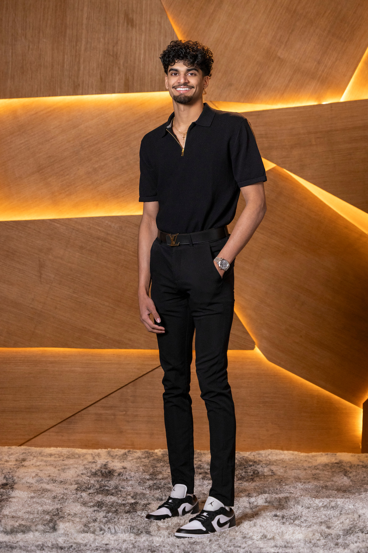 A man stands on a textured carpet in front of a wooden wall with geometric lines and warm backlighting, wearing a black shirt, black pants, and white sneakers, smiling at the camera.