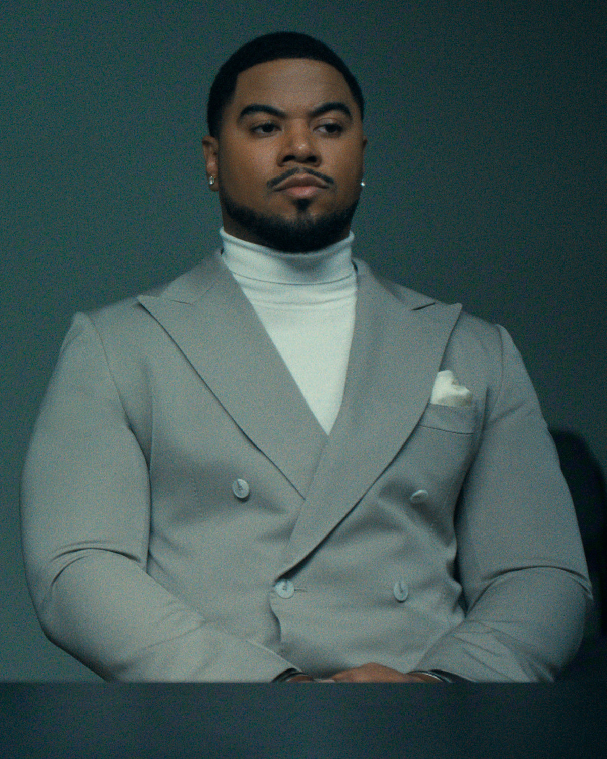 Man in a light gray double-breasted suit and white turtleneck sits at a table with a serious expression in a dimly lit, minimalist room.