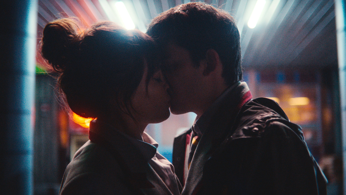 Two people kissing at night under bright neon lights, with a blurred urban background, creating a romantic and intimate atmosphere.