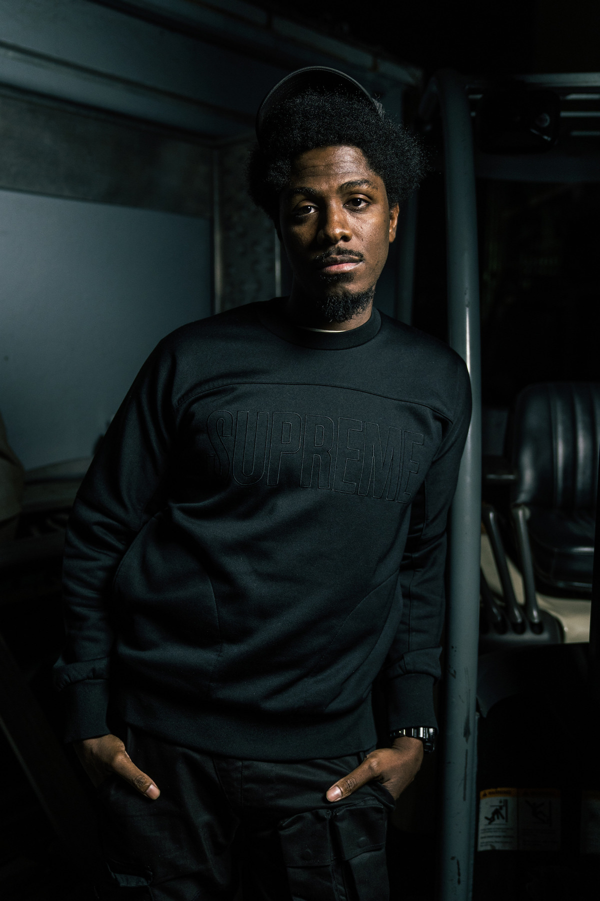 Man in black "Supreme" sweatshirt standing indoors next to industrial equipment, with his hands in his pockets, looking at the camera in a dimly lit environment.
