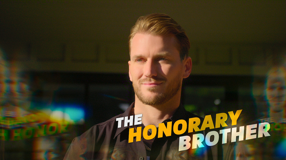 A man with light hair and beard stands outdoors, smiling in sunlight. The colorful, bold text "The Honorary Brother" is displayed across the image, with blurred reflections and a modern, vibrant atmosphere.