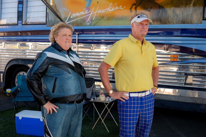 Two people stand outdoors beside a blue tour bus. One wears a blue tracksuit and the other wears a yellow polo with blue plaid pants and a visor. There are folding chairs, a cooler, and a table in the background.