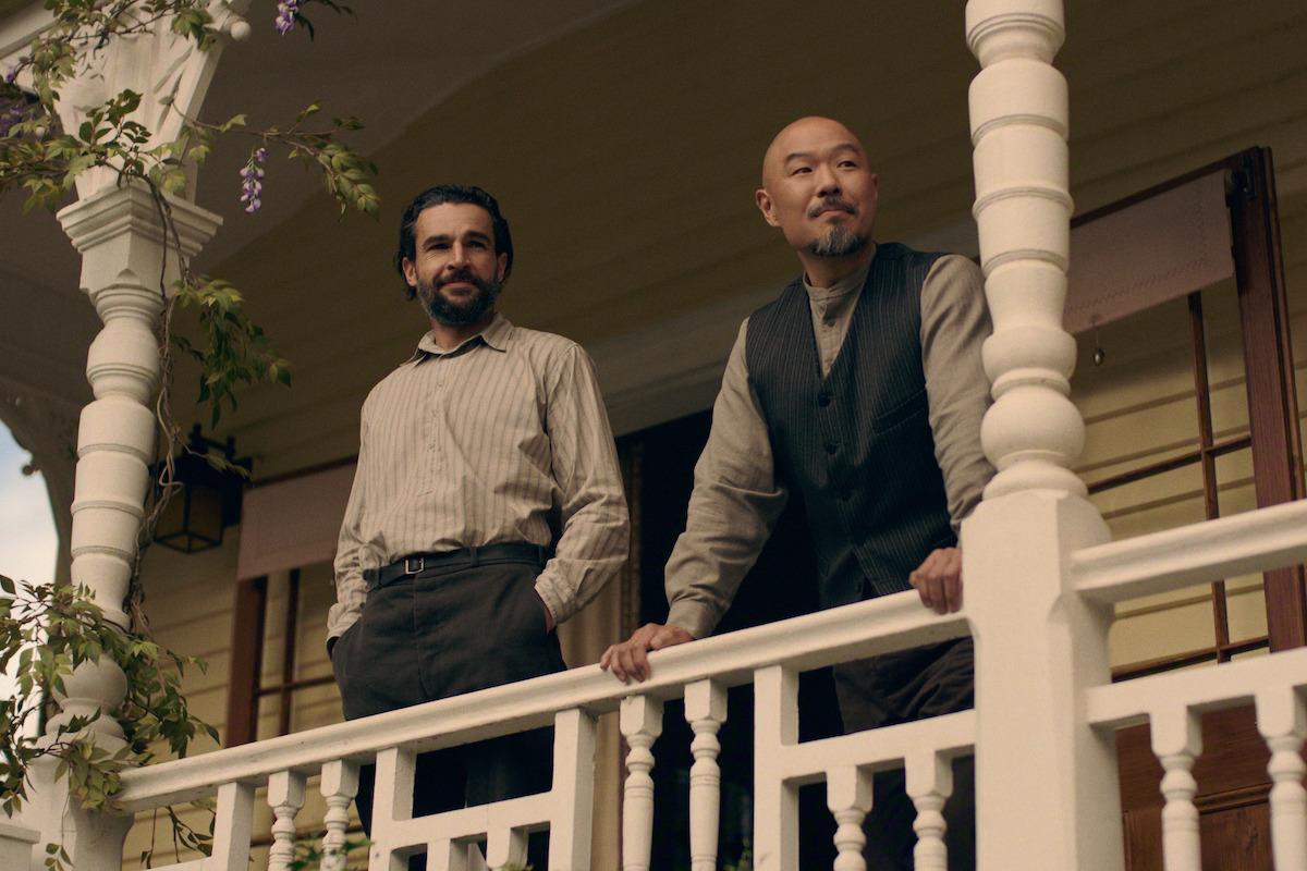 Two men stand on the porch of a house, leaning on the white railing. The setting appears to be outdoors during the day, and the porch has decorative columns with greenery visible around them.