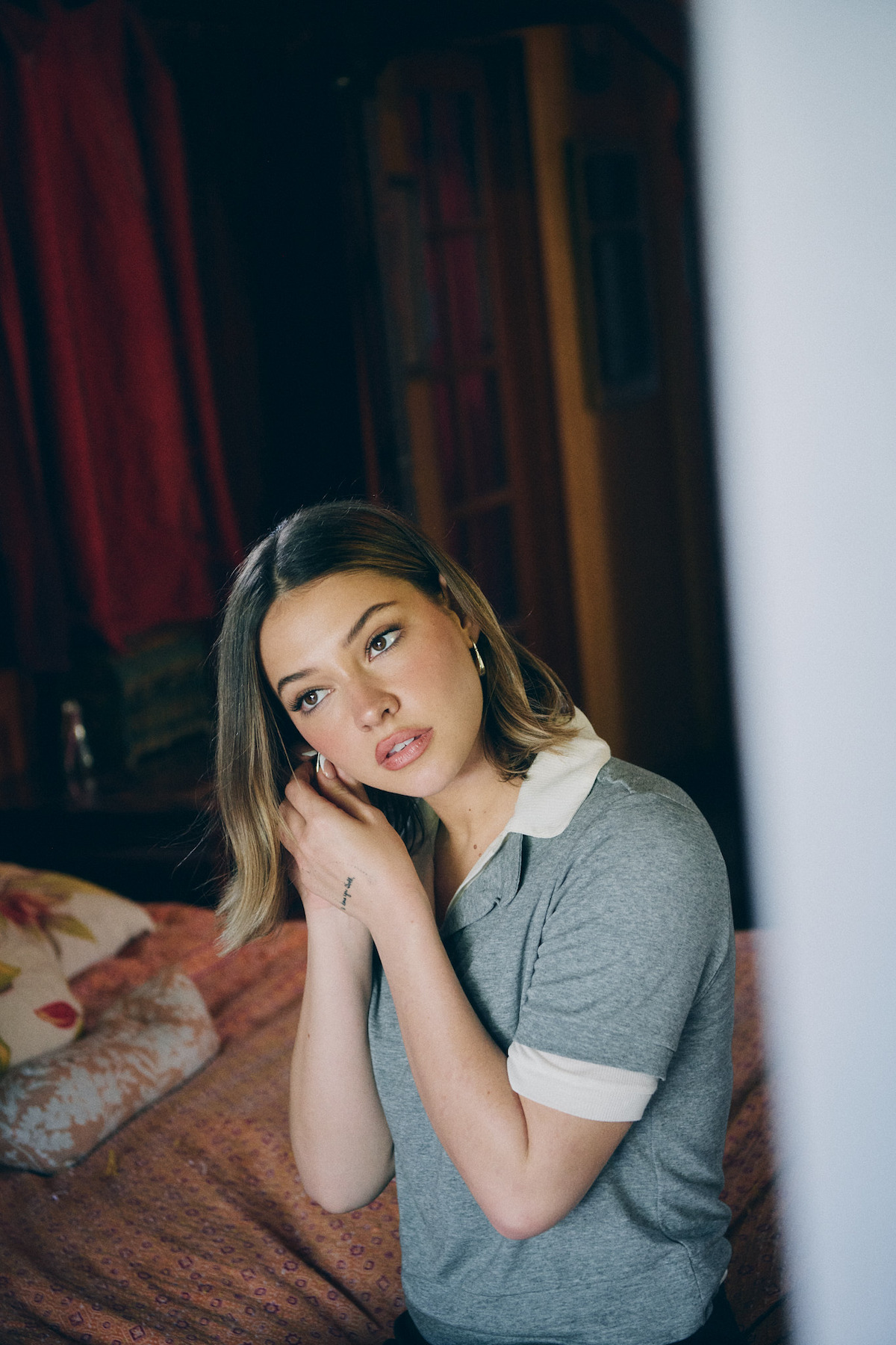 Woman in a gray collared shirt stands in a cozy bedroom with patterned bedding and red drapes, adjusting her earring and looking slightly off-camera in natural light.