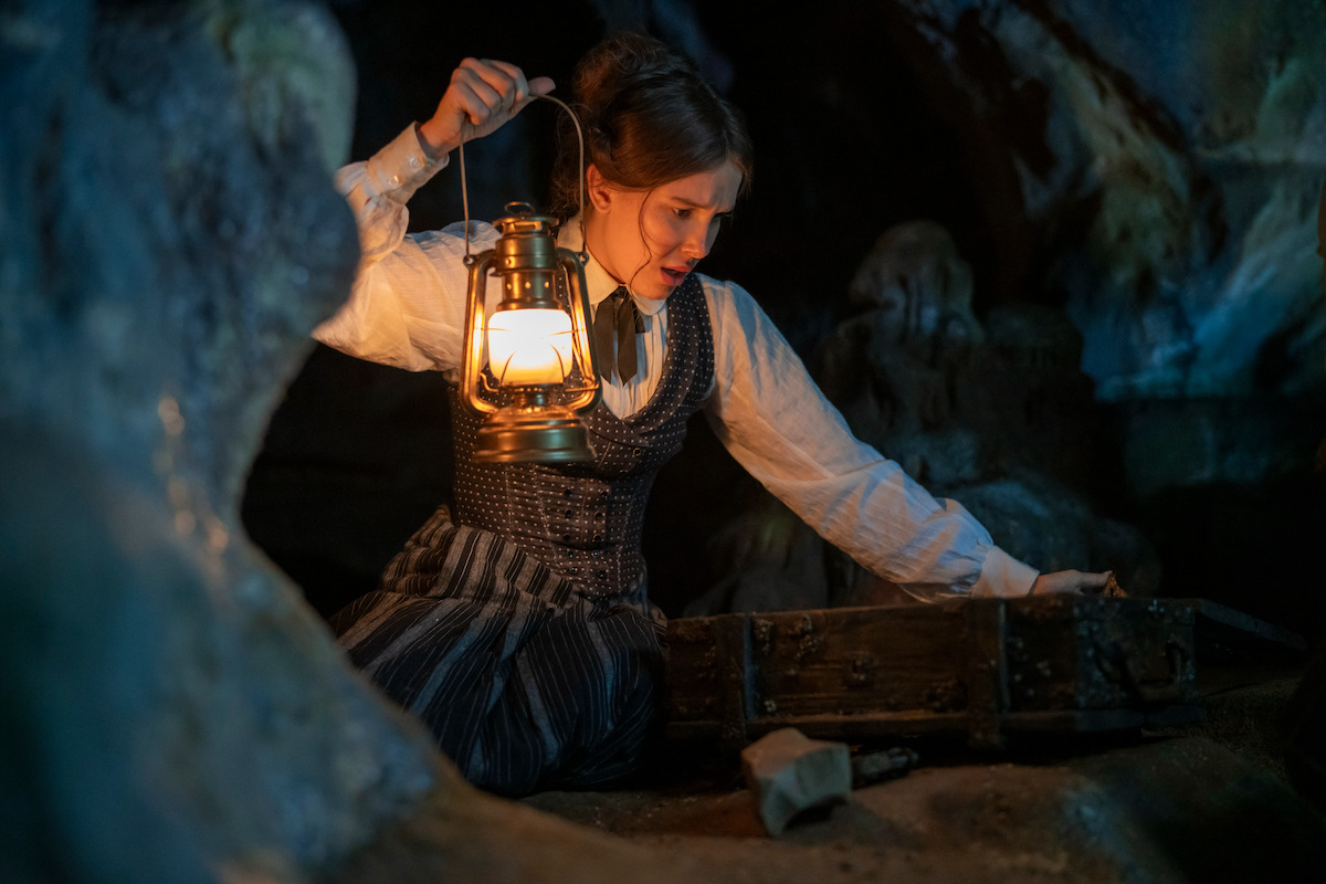 Woman holding a lantern kneels in a dark cave, illuminating an old wooden chest in front of her as she searches the contents, surrounded by rocky walls and shadows.