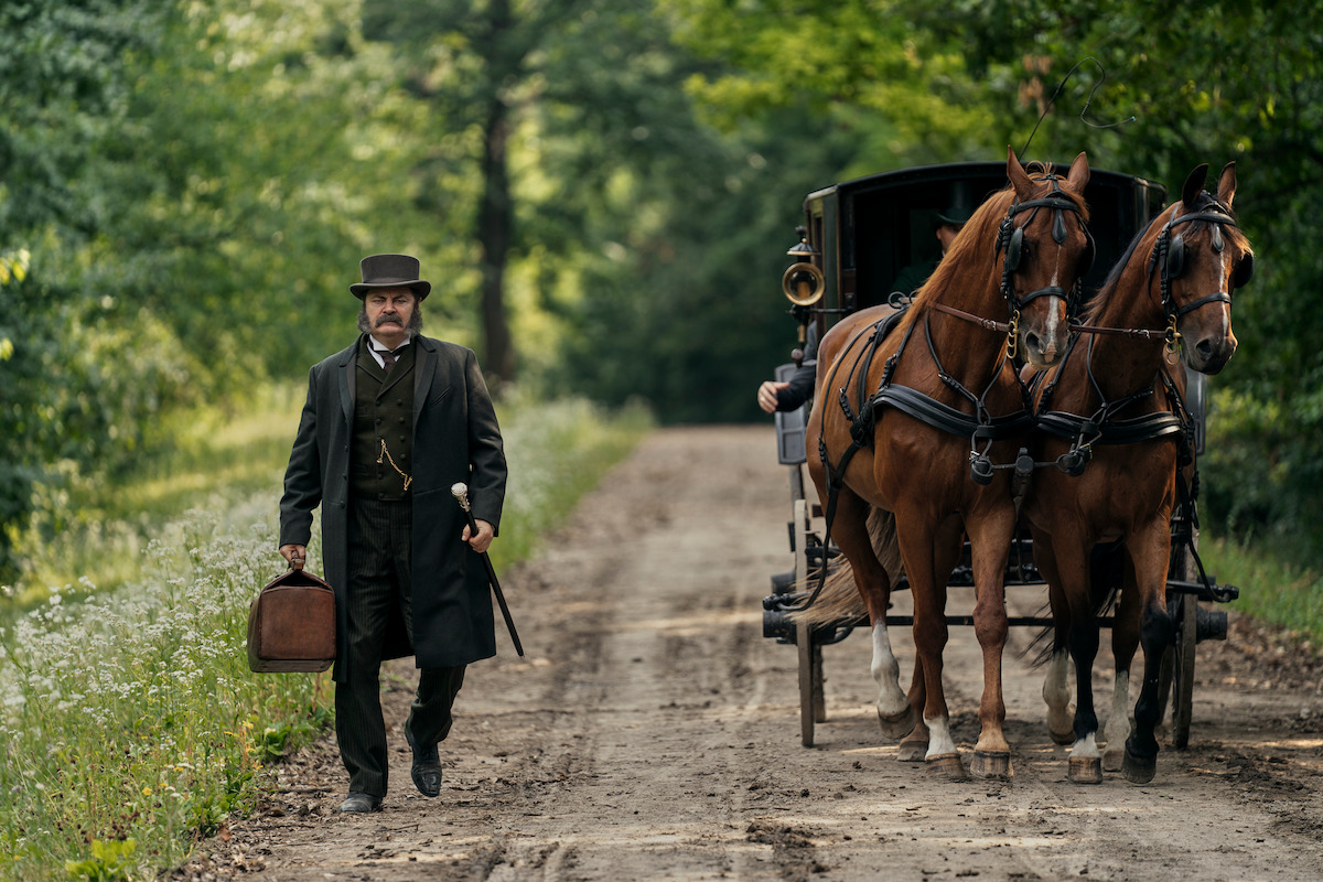 A man in period clothing with a suitcase walks on a dirt road next to a horse-drawn carriage in a lush, green forest setting, evoking a calm, historical atmosphere.