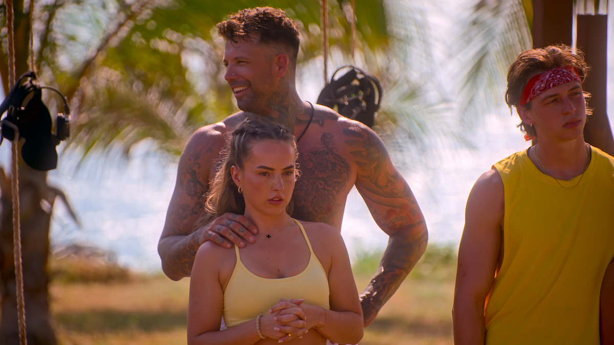 Tattooed man standing behind a woman with hands on her shoulders, another young man in yellow tank top beside them. They are outdoors, with palm trees and sunlight in a tropical beach environment.