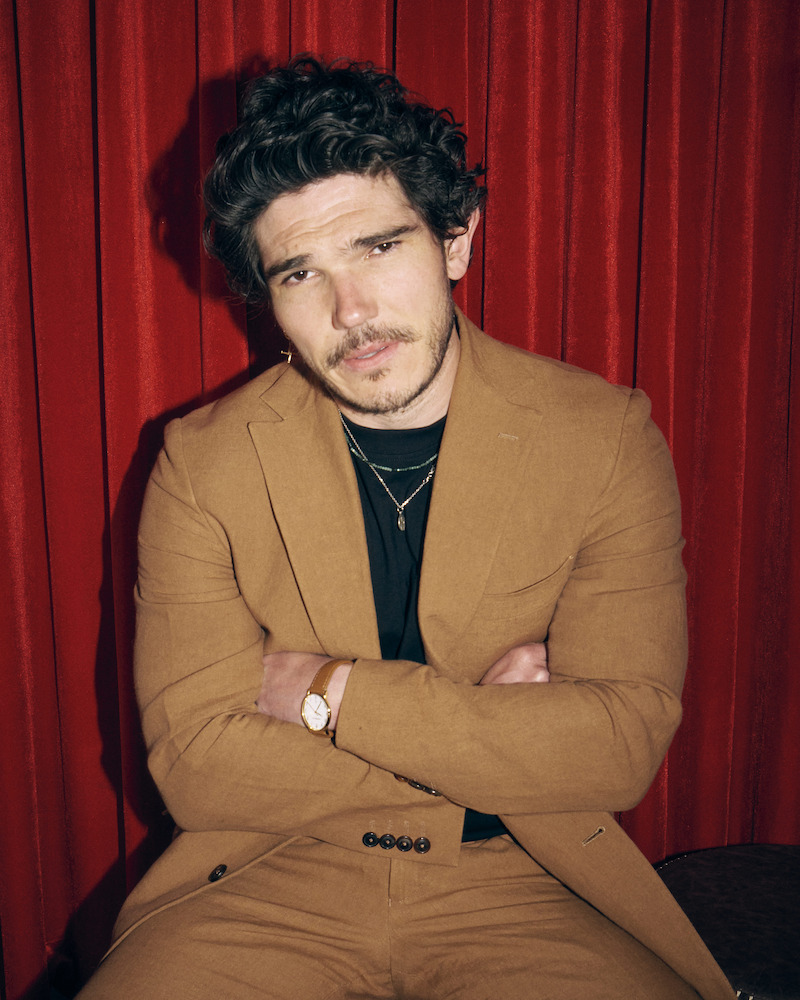 Man in light brown suit with black shirt, arms crossed, wearing necklaces and a watch, sitting in front of red curtains in a studio setting.
