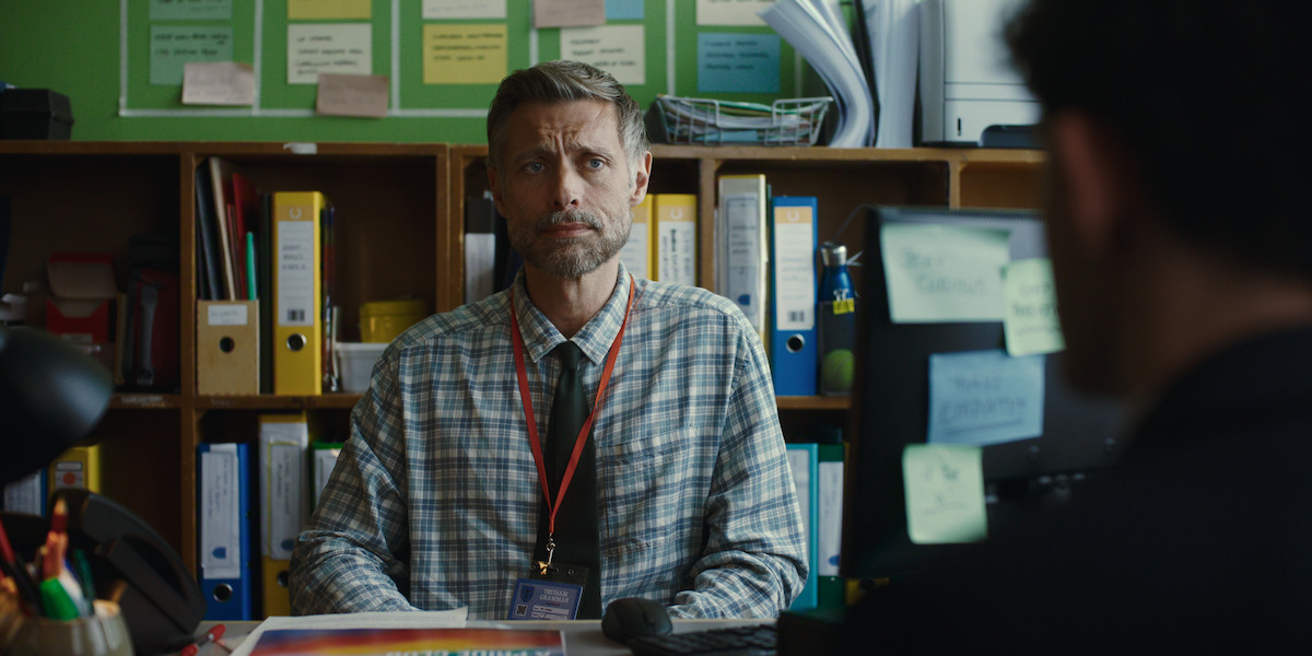 A man wearing a plaid shirt and lanyard sits at a desk in an office with shelves of binders and colorful papers behind him, while another person is seen blurred in the foreground facing him.