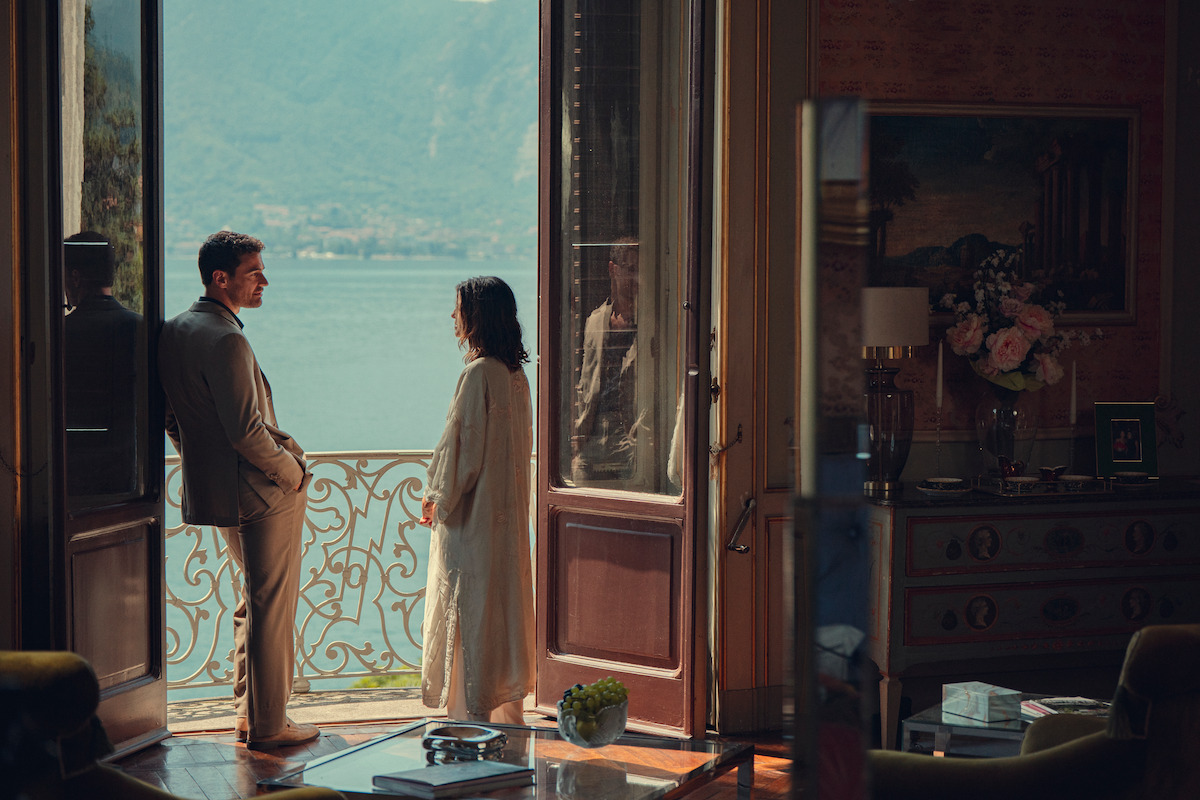 A man and woman stand on a balcony overlooking a lake, seen from inside a richly decorated room with large open doors, elegant furniture, a vase of flowers, and mountains visible in the background.