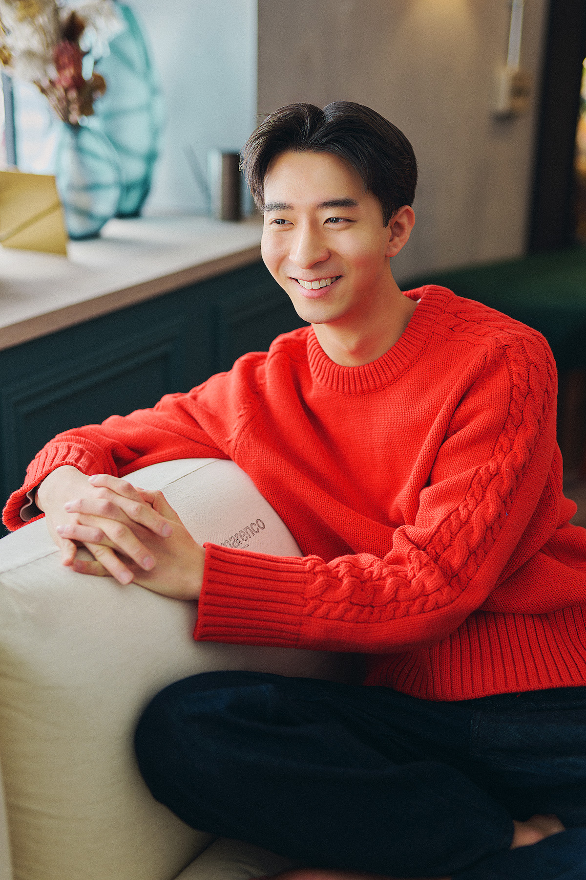 Young man in a bright red sweater sits casually on a couch in a cozy, modern living room, smiling warmly. The atmosphere is relaxed and inviting with natural light and tasteful decor in the background.
