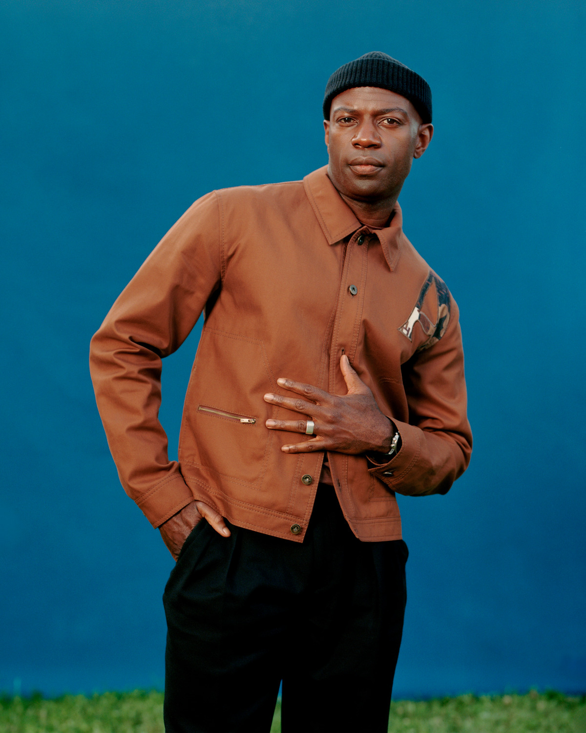 David Gyasi, wearing a black hat and a brown jacket with black pants, standing in front of a blue backdrop.