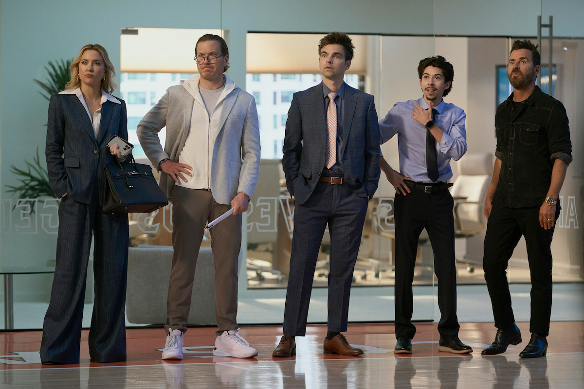Five people in business attire stand indoors on a basketball court in a modern office, looking focused and attentive, possibly watching or listening to something off camera. The background shows glass walls and office furniture.