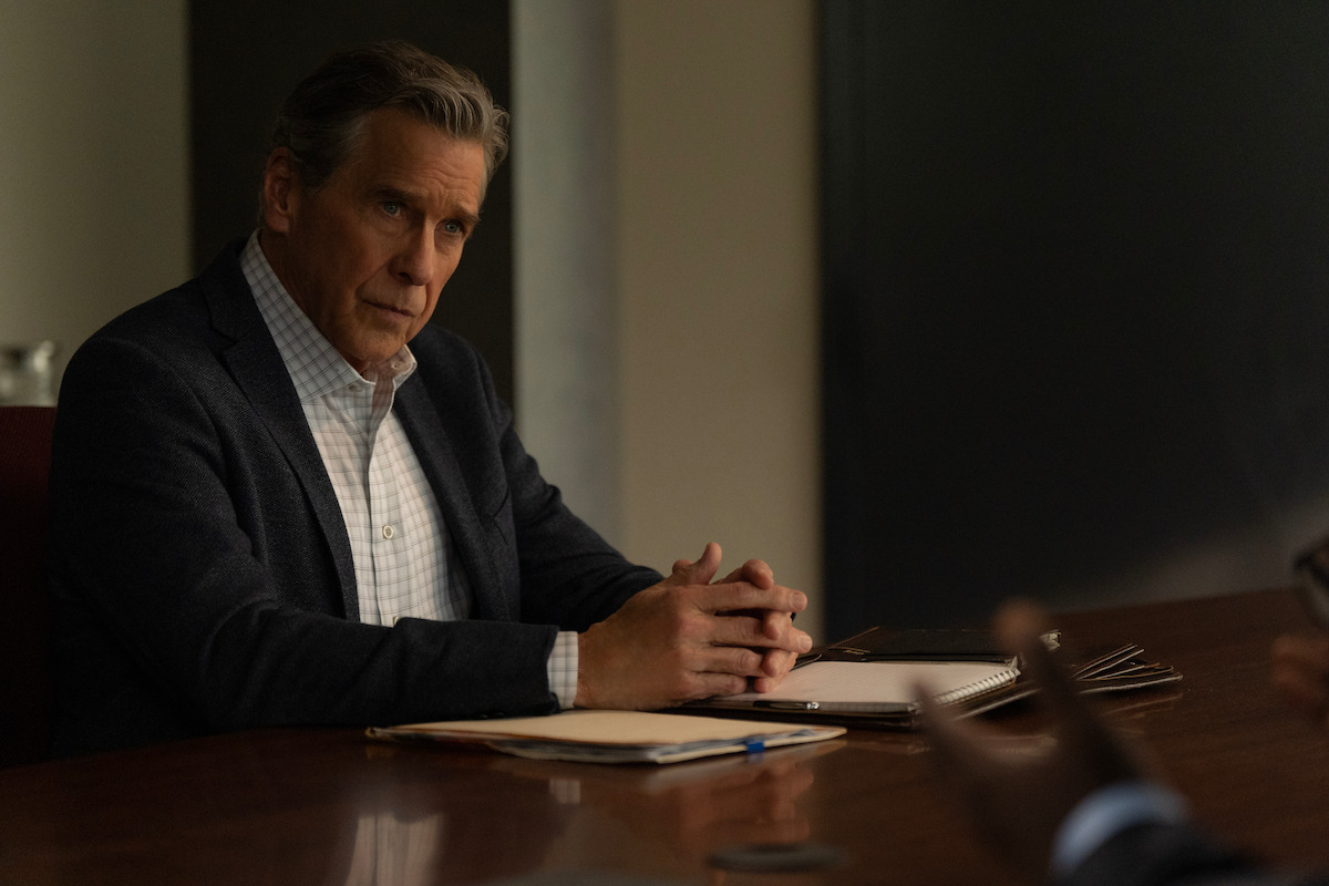 A man in a suit sits at a conference table with folders and papers, listening intently in a dimly lit office or meeting room setting.