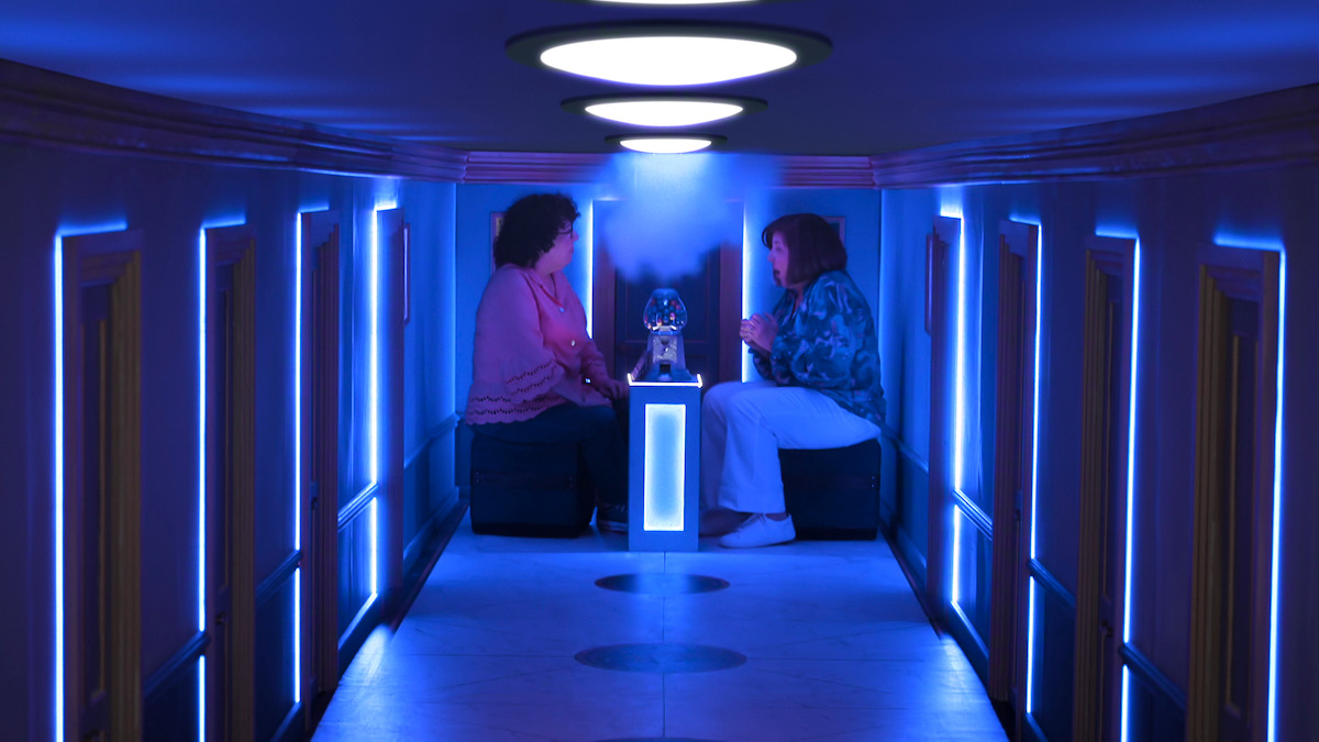 Two people sit facing each other in a narrow, futuristic hallway with blue neon lighting, talking across a small table with a lit object between them. The corridor has a modern, sci-fi ambiance.