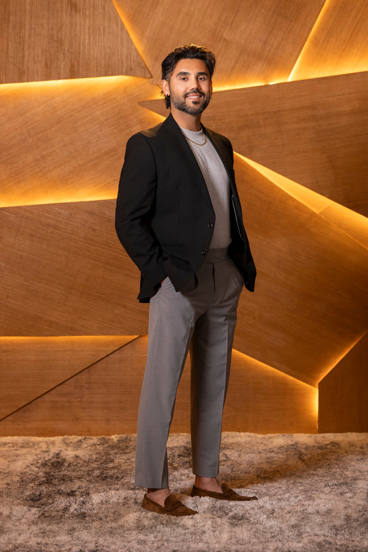 A man stands on a carpeted floor in front of a modern wooden wall with geometric panels and warm lighting, wearing a blazer, t-shirt, trousers, and loafers, hands in pockets, looking at the camera and smiling.
